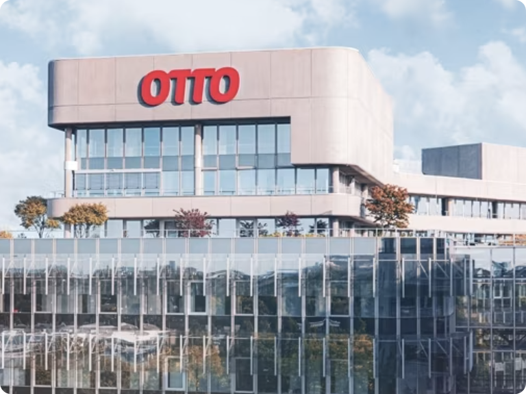 The image shows a modern office building with a large sign on the top that reads "OTTO" in red letters. The building has multiple floors with large glass windows and some greenery, including trees, on the rooftop. The sky is clear with some clouds, providing a bright and pleasant backdrop. This image is interesting as it depicts the headquarters of a company named OTTO, showcasing its contemporary architecture and environmentally friendly design elements such as rooftop greenery.